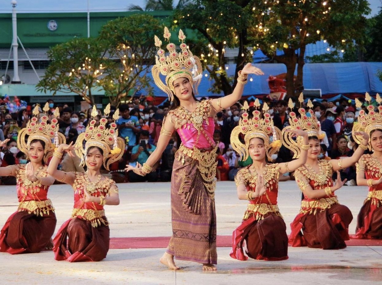 Eine Gruppe von Frauen in traditioneller thailändischer Kleidung, die vor einem sitzenden und stehenden Publikum tanzen, mit Bäumen, Lichtern, Pfählen, einem Gebäude und einem klaren blauen Himmel im Hintergrund.
