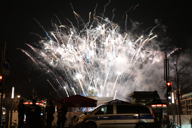 Polizeiauto vor einer Menschenmenge bei einer Silvesterfeier in Berlin geparkt, mit farbigen Feuerwerken, die den Himmel und die Gebäude im Hintergrund erhellen.
