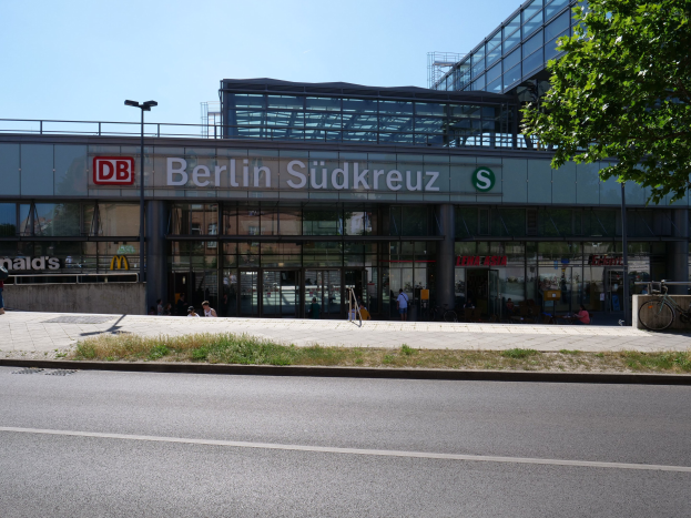 Großes Glaswand-Gebäude des Berliner Südkreuz-Bahnhofs mit Text an seiner Fassade, umgeben von Straßeninfrastruktur, Fahrzeugen, Fußgängern, Fahrrädern, Bäumen und einem klaren Himmel.