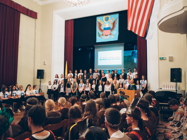 Eine Gruppe von Menschen steht auf einem Podium mit einem Mikrofon, flankiert von Fahnen und Lautsprechern, und spricht vor einem sitzenden Publikum während eines kulturellen Ereignisses.