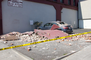 Eine zerbrochene Ziegelwand liegt auf dem Boden mit verstreuten roten Ziegeln, einem geparkten Auto, einer Topfpflanze und einem Gebäude mit einer 'Kautionsfreiheit'-Plakette im Hintergrund.