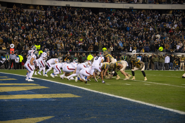 Ein Fußballspiel im Gange mit Spielern auf dem Rasen in Sportkleidung und Helmen, aus der Perspektive eines Stadionplatzes mit Zuschauern und Werbetafeln im Hintergrund.