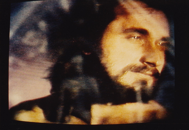 Mann mit dunklem Bart und Schnurrbart, ernster Gesichtsausdruck, trägt ein weißes Hemd, auf einem Fernseher mit dunklem Hintergrund.