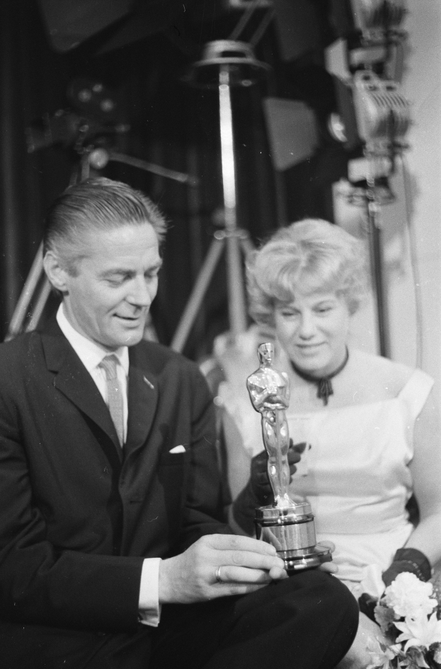 Ein Mann in einem schwarzen Blazer, weißem Hemd und Krawatte hält eine Oscar-Statue in der Hand und sitzt neben einer Frau, die einen Blumenstrauß hält, beide in formeller Kleidung, mit abgedunkeltem Hintergrund in Schwarz-Weiß.