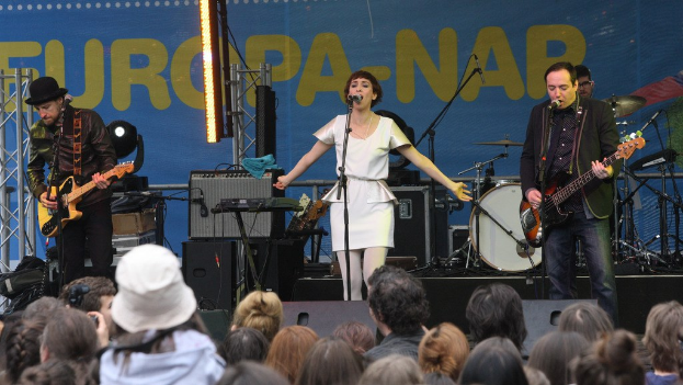 Vier Musiker auf der Bühne: Eine Frau in einem weißen Kleid singt in der Mitte, flankiert von zwei Männern mit Gitarren, vor einem Publikum und Lautsprechern an der Wand im Hintergrund.