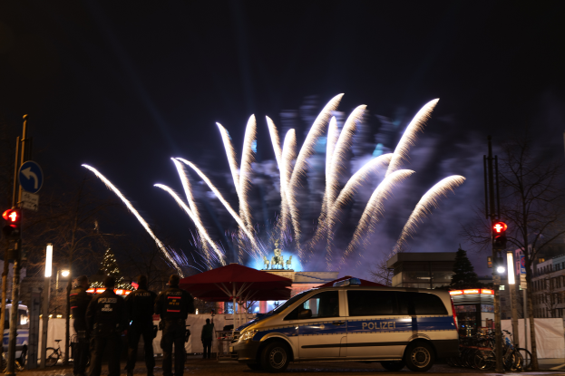 Ein Polizeiwagen steht vor einer Menschenmenge, mit beleuchteten Gebäuden und Bäumen im Hintergrund, während farbige Feuerwerke den Himmel erhellen.