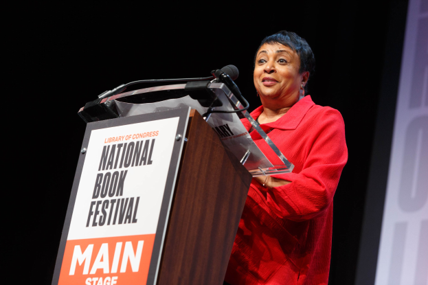 Frau in einem roten Kleid, die an einem Pult spricht, das mit "National Book Festival" beschriftet ist, mit einem Bildschirm rechts daneben.