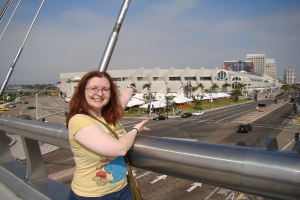 Eine Frau steht lachend auf einer Brücke, mit Autos auf der Straße darunter und Bäumen und Gebäuden im Hintergrund.
