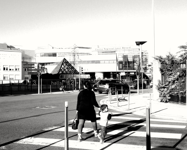 Eine Frau in einem schwarzen Kleid und ein Kind gehen gemeinsam über eine Fußgängerüberquerung, mit Fahrzeugen, Pfosten, Bäumen, Gebäuden und einem klaren Himmel im Hintergrund.