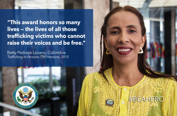Eine Frau in einem gelben Kleid lächelt in die Kamera, mit einem Logo und Text auf der linken Seite, die sie als Betty Pedraza Lozano, Kolumbien - TIP-Heldin 2015 identifizieren, vor einem unscharfen Hintergrund von Säulen, Pflanzen und anderen Gegenständen.