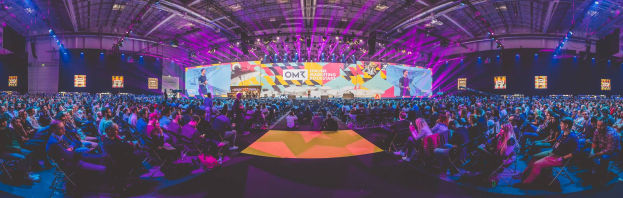 Eine große Menschenmenge, die auf Stühlen sitzt und einer hell erleuchteten Bühne mit mehreren stehenden Personen gegenüber, wahrscheinlich auf einem Online-Marketing-Rockstars-Event, umgeben von Bildschirmen.