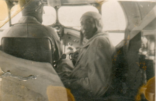 Schwarzes und weißes Foto von zwei Männern in einem Cockpit, einer sitzt im Pilotensitz und trägt eine Uniform, der andere steht neben ihm und hat einen ernsten Gesichtsausdruck.