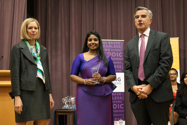 Zwei Frauen und ein Mann stehen auf einer B├╝hne mit einem braunen Vorhang dahinter; die Frau in der Mitte h├Ąlt einen Preis, und mehrere Preise sind auf dem Tisch vor ihnen.