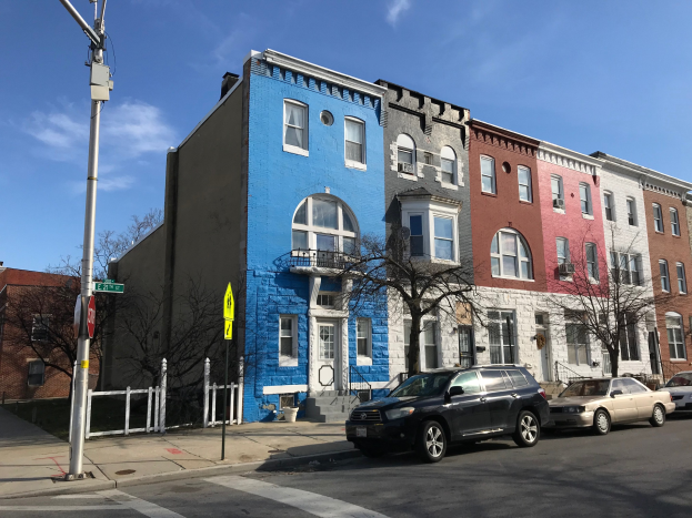 Eine Reihe bunter Häuser mit geparkten Autos auf einer Straße, die von Bäumen, Pfählen, Brettern und einem Zaun gesäumt ist, unter einem bewölkten Himmel in Baltimore, Maryland.
