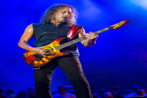 Eine Person in einem schwarzen Kleid steht in der Mitte eines musikalischen Konzerts und spielt eine Gitarre, während das Publikum von unten zusieht.