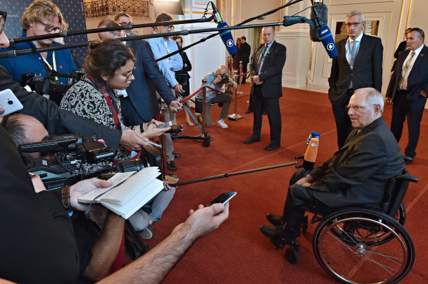 Ein Mann im Rollstuhl umgeben von Reportern mit Kameras, Handys und Mikrofonen sowie einem Banner, Scheinwerfern und einer Wand im Hintergrund.