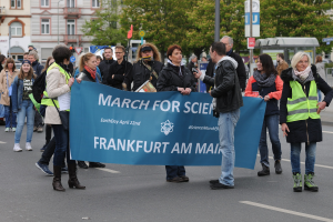 Eine bunte Gruppe von Menschen marschiert die Straße entlang, trägt ein "March for Science Frankfurt am Main"-Schild, mit Bäumen, Polen, Schildern, Gebäuden und einem klaren blauen Himmel im Hintergrund.
