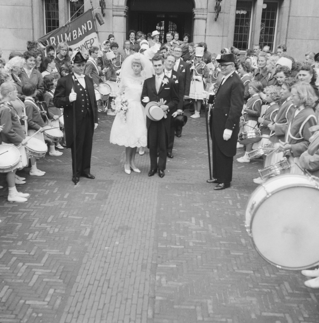 Schwarzes und weißes Foto einer Braut in einem weißen Kleid und Schleier und Bräutigam mit einem Blumenstrauß, der eine Straße entlang geht, vorbei an einer Menge, einige halten Musikinstrumente, mit einem Banner und Gebäude im Hintergrund.