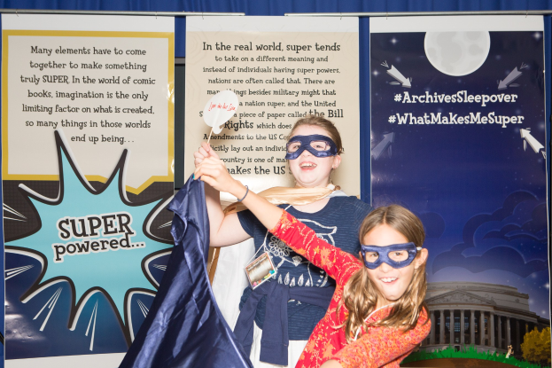 Zwei junge Mädchen in Superheldenkostümen posieren vor einem blauen Vorhang bei einem Schulereignis, eine hält ein Tuch und beide tragen Masken.