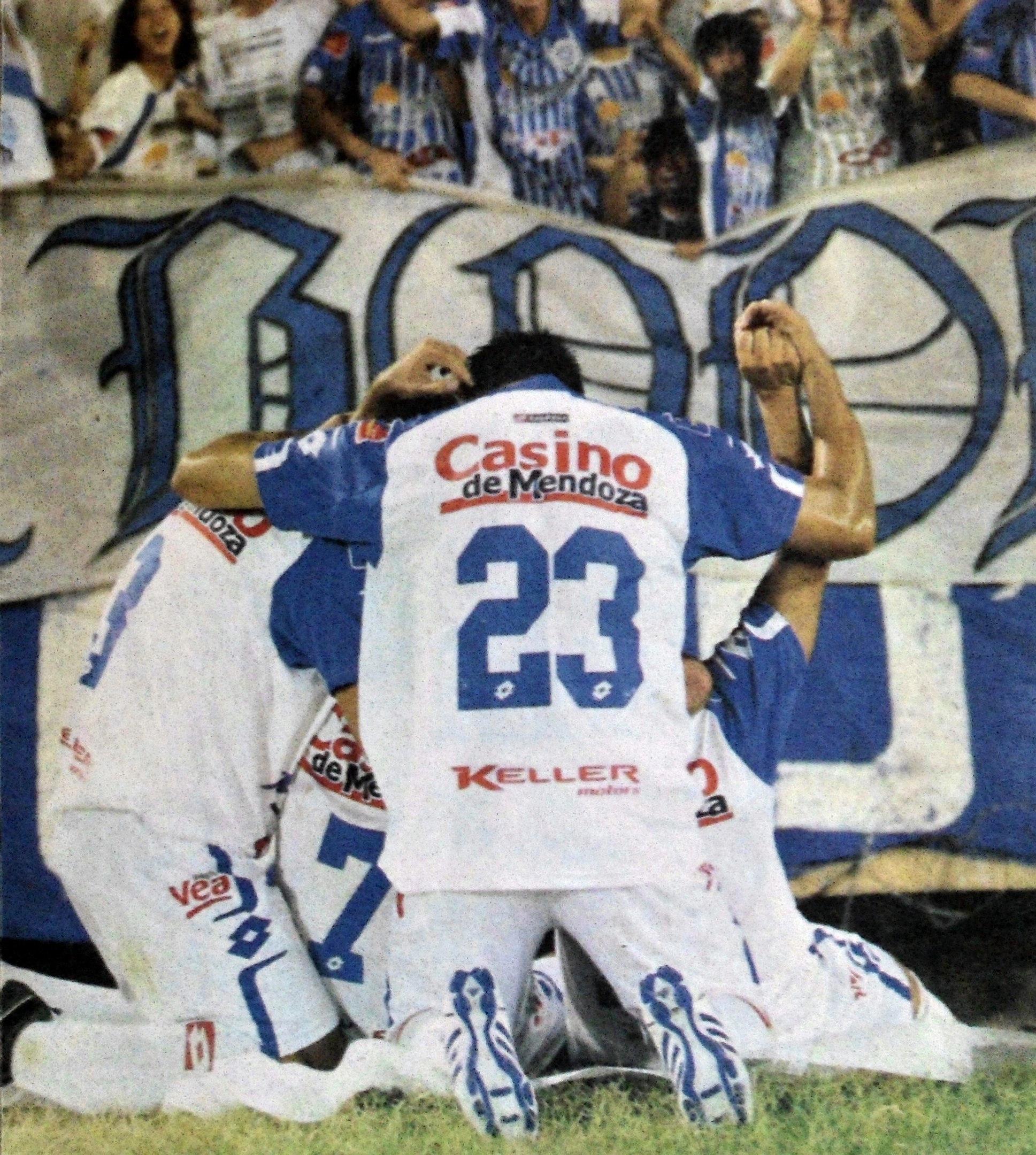 Eine Gruppe von Fußballspielern, die ein Tor feiern, auf dem Boden vor einer jubelnden Menge, mit einem Banner mit Text im Hintergrund.