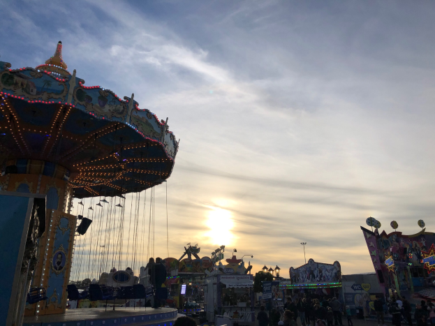 Ein Karussell auf einem Rummelplatz mit Menschen darauf, beleuchtet von der untergehenden Sonne, umgeben von Fahrgeschäften, Pfählen und Lichtern, mit einem bewölkten Himmel im Hintergrund.