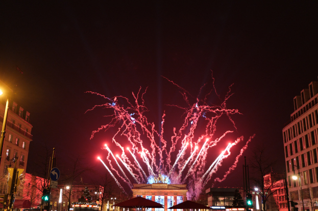 Eine belebte Straßenkreuzung in Berlin an Silvester, voller Menschen, Fahrzeuge und beleuchtet von Gebäudelichtern und Feuerwerk am Himmel.