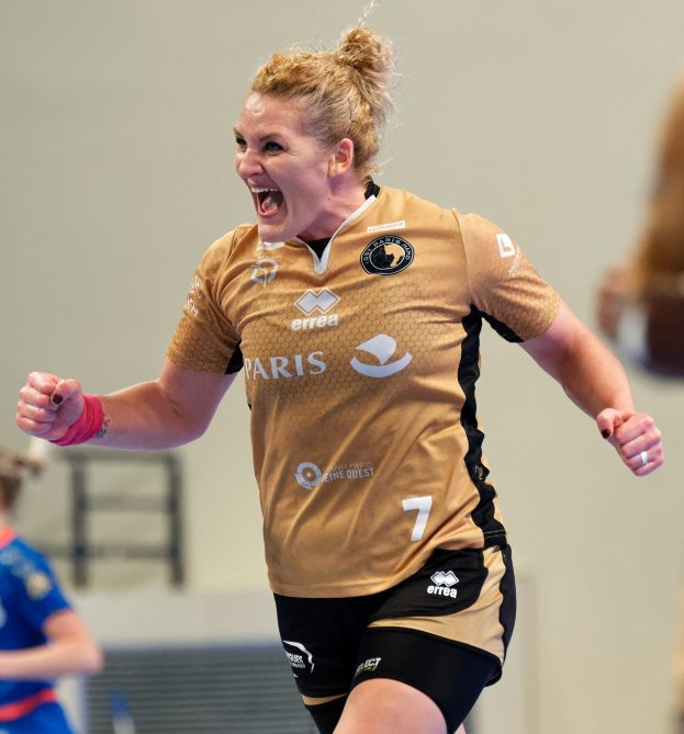 Eine Frau in braunem T-Shirt und schwarzen Shorts läuft triumphierend auf einem Futsalplatz, mit zwei anderen Frauen im Hintergrund.