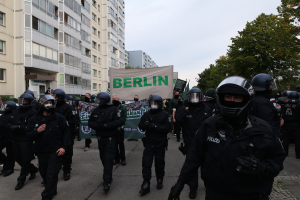 Eine Gruppe von Polizisten in Schutzausrüstung, Helmen und mit Schildern geht eine Straße entlang mit Gebäuden, Bäumen und einem klaren blauen Himmel im Hintergrund.