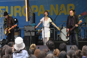 Ein Konzertauftritt mit vier Personen auf der Bühne: Eine Frau in einem weißen Kleid singt in der Mitte, zwei Männer spielen Gitarren links und rechts daneben und eine Menge im Vordergrund mit einer Wand und Lautsprechern im Hintergrund.