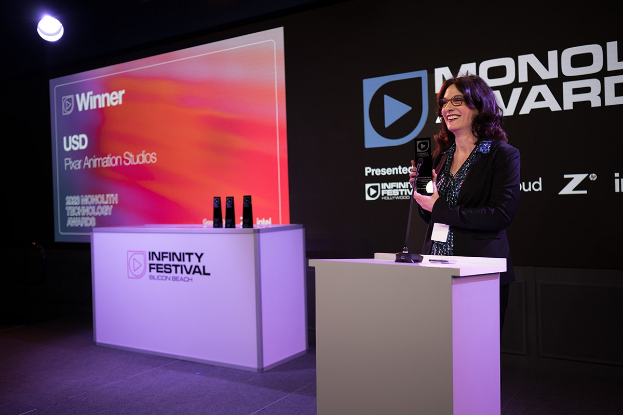Frau in einem schwarzen Kleid, die vor einem Podium steht und einen Preis hält, mit einem Mikrofon und anderen Gegenständen auf dem Podium und einem Werbeplakat im Hintergrund.